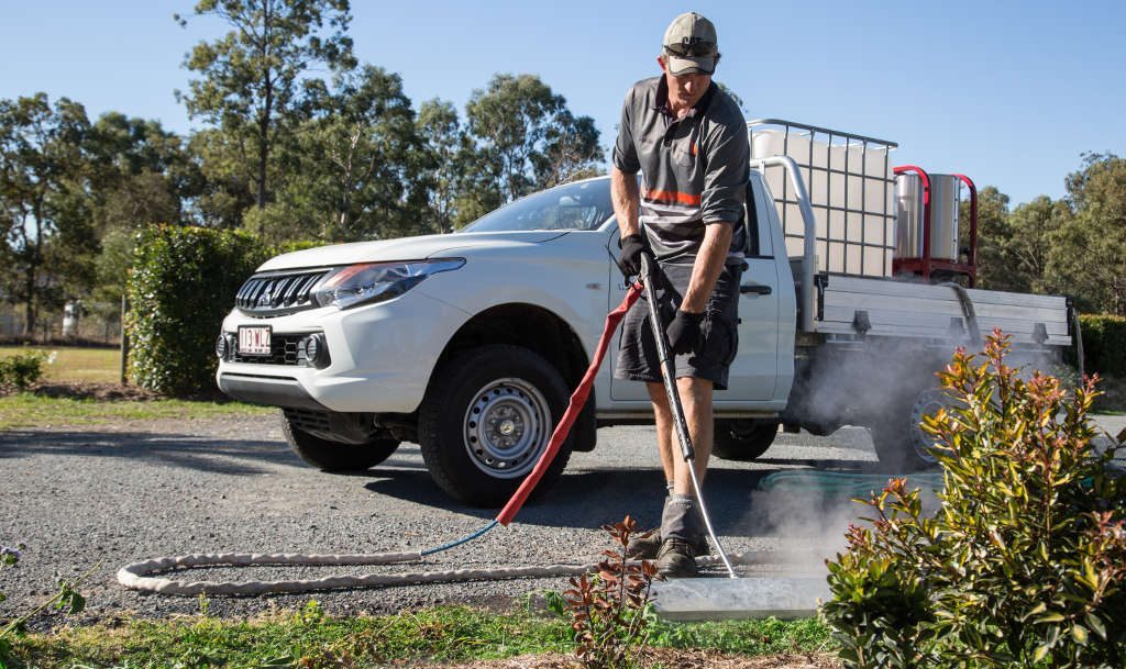 Worker using a steam weed wand on a gravel driveway beside a white pickup with a water tank