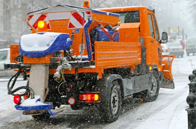 Orange salt spreader truck with a spinner on the rear driving through a snowstorm
