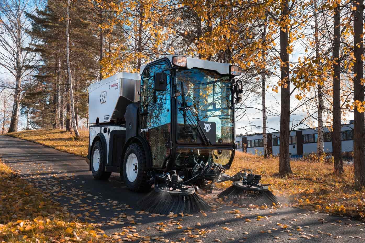 Multihog street sweeper with twin side brushes sweeping a path lined with fall foliage
