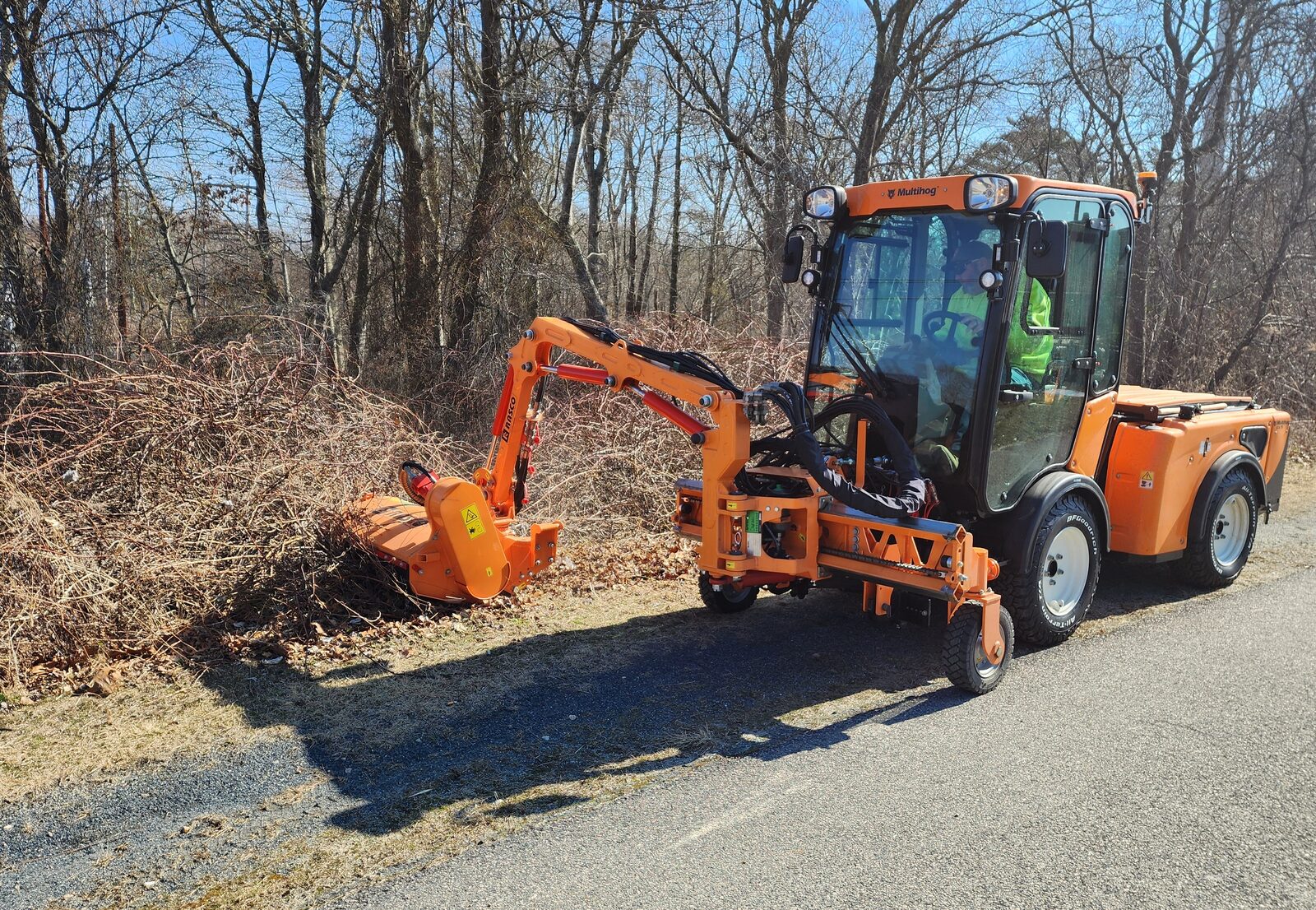 Orange Multihog tractor with boom-arm flail mower cutting brush along a paved trail, operator in cab