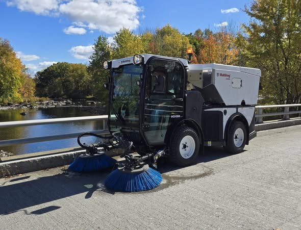 Multihog CV300 street sweeper with blue side brushes on a bridge next to a river