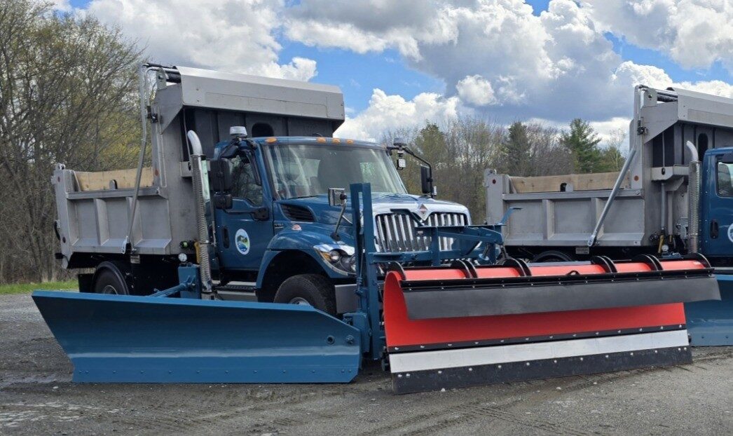 Blue municipal dump truck with blue front plow and red and white wing plow, second truck behind