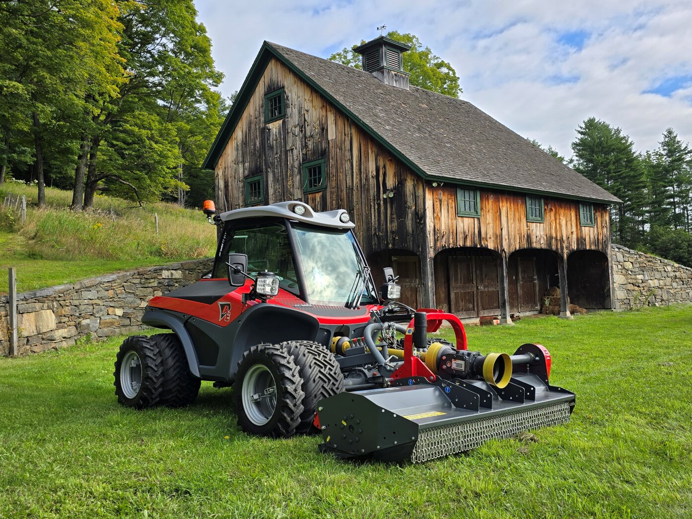 Red and black Aebi tractor with rear flail mower parked on grass beside a rustic barn